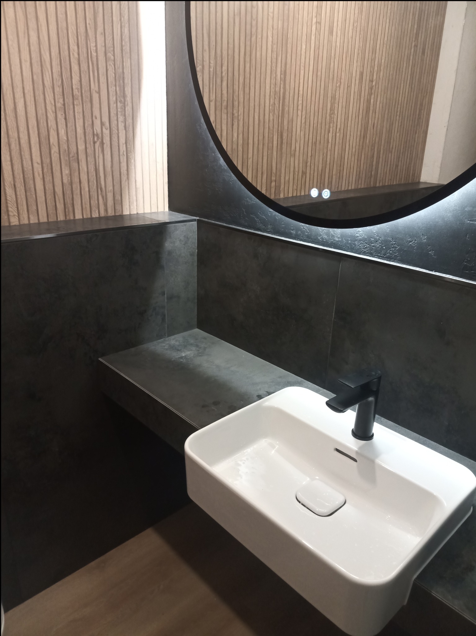 Nowoczesna łazienka z umywalką nablatową, czarną armaturą i podświetlanym lustrem w okrągłej ramie na tle ściany z drewnopodobnych paneli.
