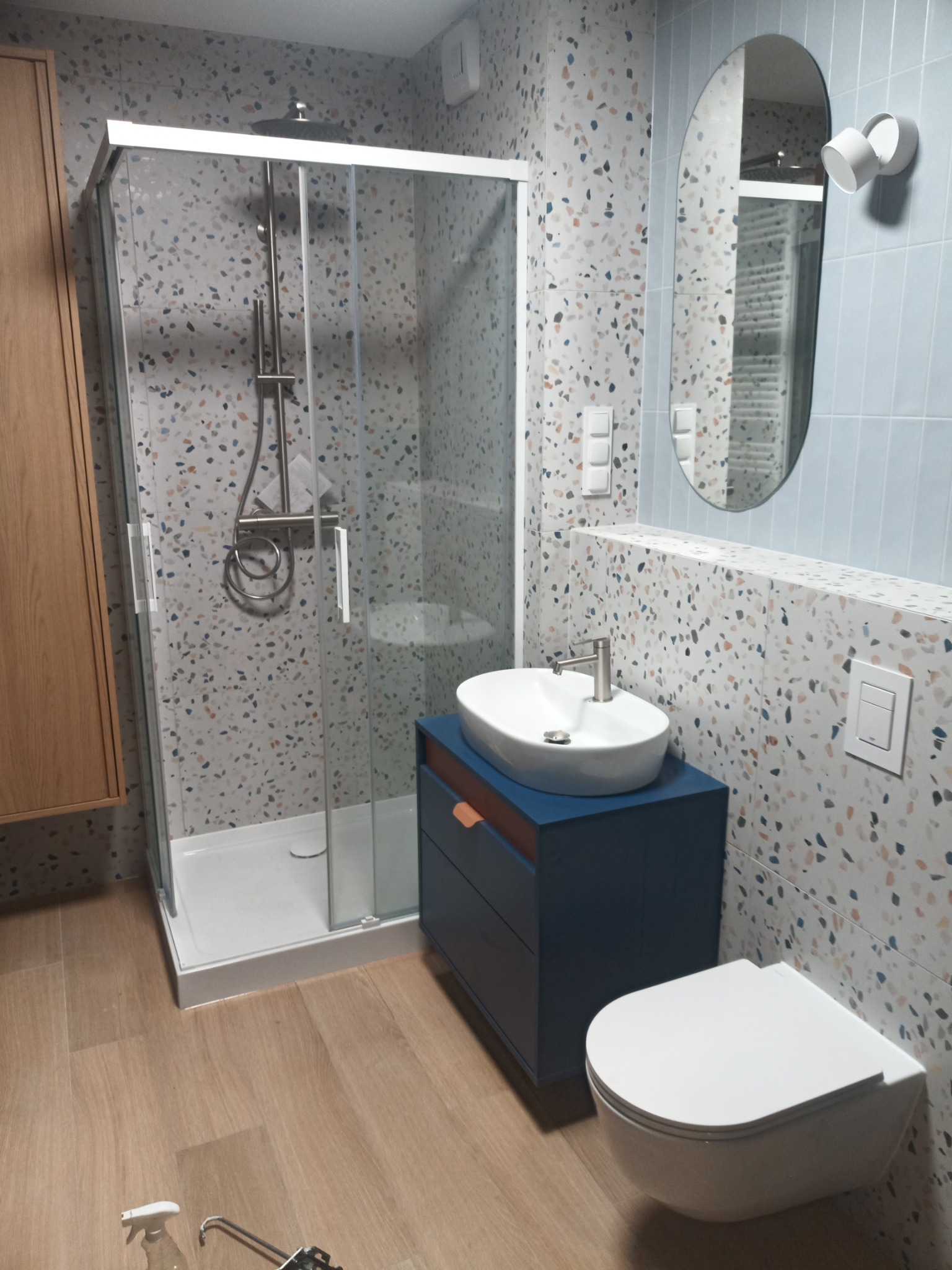 Nowoczesna łazienka z kabiną prysznicową, umywalką nablatową na niebieskiej szafce, wiszącą toaletą i lustrem o nieregularnym kształcie. Ściany wyłożone płytkami imitującymi lastryko, podłoga...