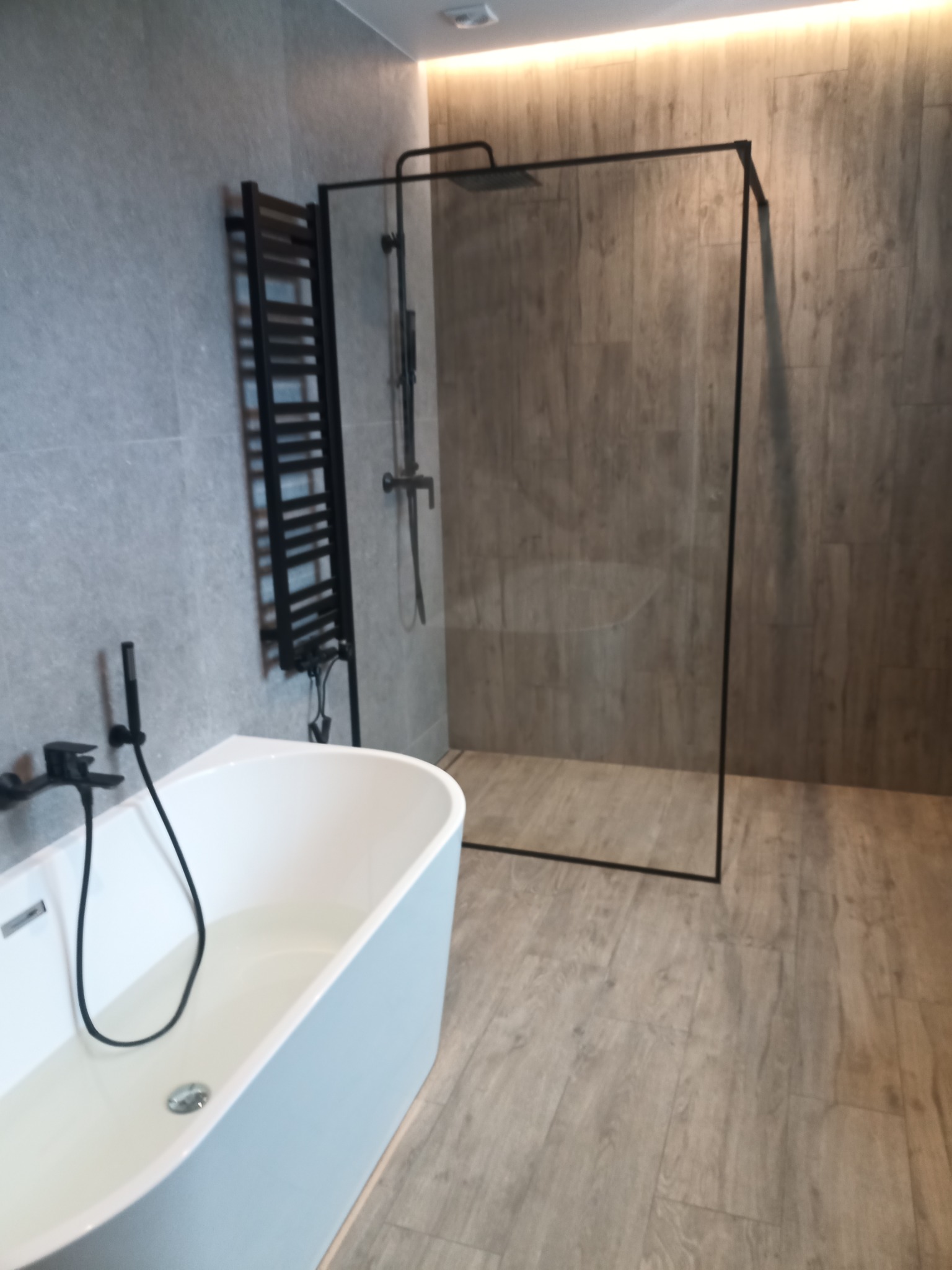 Nowoczesna łazienka z wolnostojącą wanną wypełnioną wodą, czarną armaturą i kabiną prysznicową typu walk-in z czarnymi profilami, na podłodze panele imitujące drewno, oświetlenie LED podsufitowe.