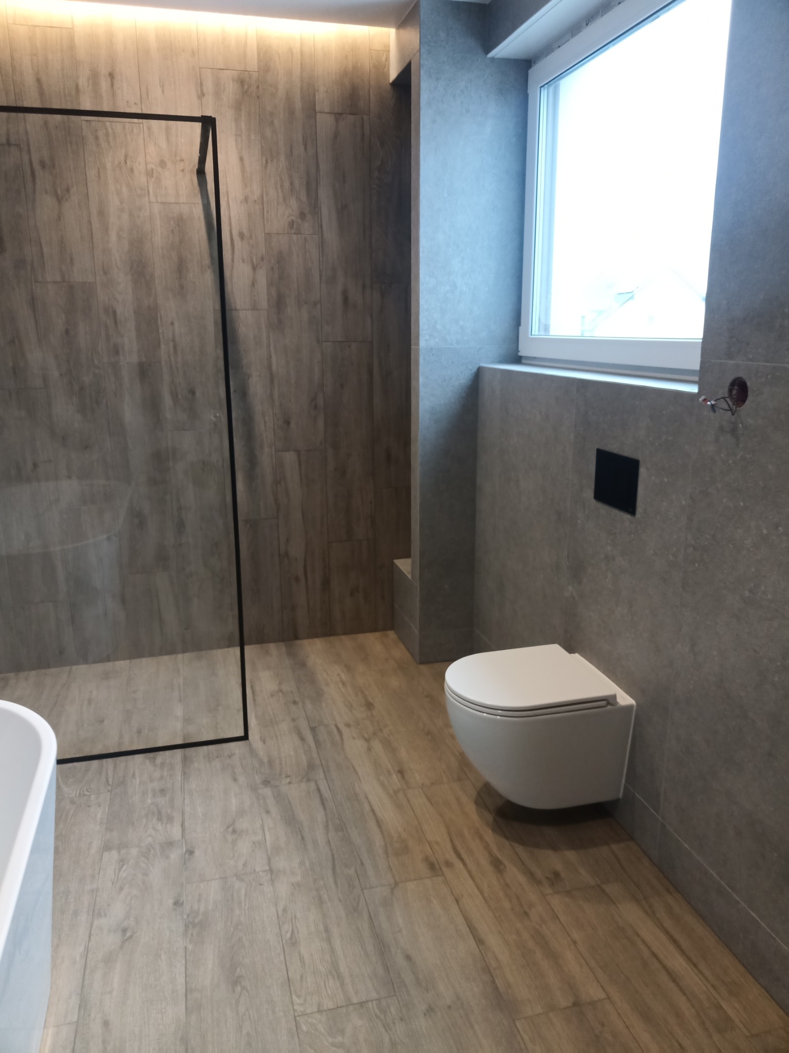 Nowoczesna łazienka z kabiną prysznicową typu walk-in, podwieszaną toaletą i płytkami imitującymi drewno na podłodze oraz szarymi płytkami na ścianach, widoczne niedokończone podłączenie...