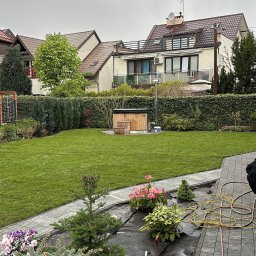 ZIELONA MAGIA MACIEJ STYCZYŃSKI - Widok ogrodu z trawnikiem, drewnianą balią z kominem i przygotowanymi sadzonkami w doniczkach na czarnej folii, w tle zabudowa jednorodzinna.