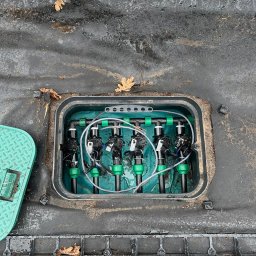 ZIELONA MAGIA MACIEJ STYCZYŃSKI - Otwarta skrzynka z zaworami i rurami systemu nawadniania, widoczne elementy sterujące i przyłącza, na zewnątrz czarna agrowłóknina i opadłe liście.