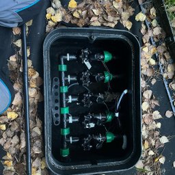 ZIELONA MAGIA MACIEJ STYCZYŃSKI - Czarny plastikowy zawór sterujący z zielonymi elementami w czarnej skrzynce w ziemi, otoczonej opadłymi liśćmi.