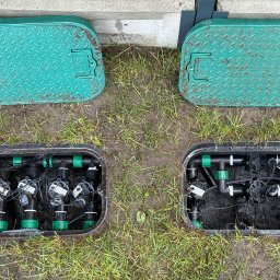 ZIELONA MAGIA MACIEJ STYCZYŃSKI - Dwa zielone, żebrowane włazy skrywające zawory nawadniające, umieszczone w ziemi z trawą. Widoczne czarne rury i złączki systemu.