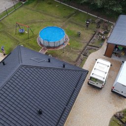 ZIELONA MAGIA MACIEJ STYCZYŃSKI - Widok z góry na dom z czarnym dachem, garaż, basen z niebieskim przykryciem, plac zabaw i dwa białe busy na posesji w trakcie prac ziemnych.