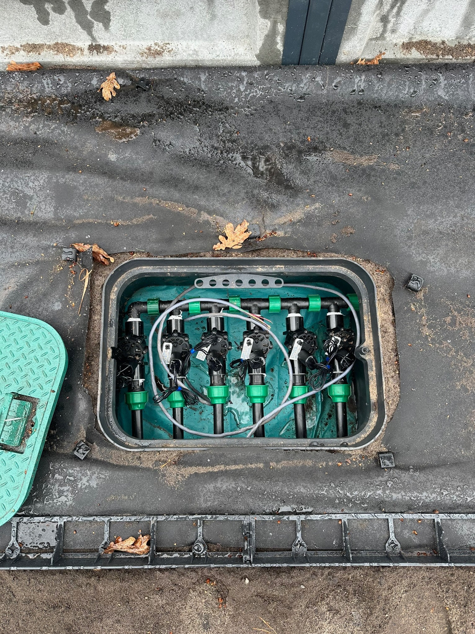 Otwarta skrzynka z zaworami i rurami systemu nawadniania, widoczne elementy sterujące i przyłącza, na zewnątrz czarna agrowłóknina i opadłe liście.