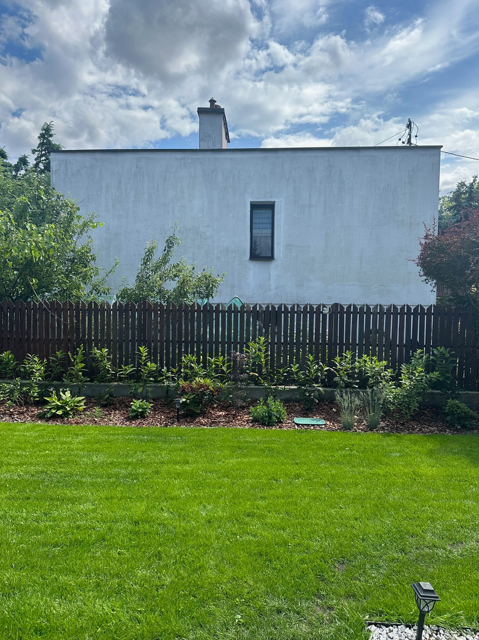 Zadbany trawnik przed domem z drewnianym ogrodzeniem i rabatą z roślinami, w tle biały budynek z jednym oknem i kominem.