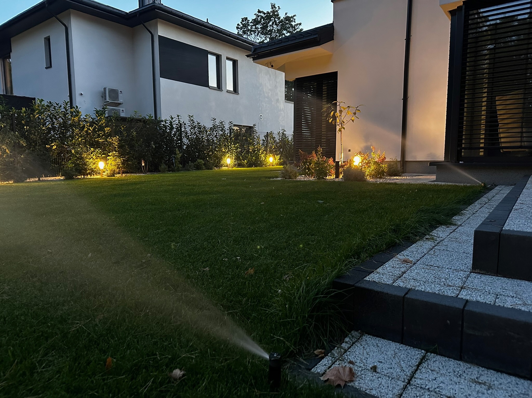 Nowoczesny ogród z automatycznym systemem nawadniania trawnika, oświetleniem punktowym i kostką brukową przy domu.