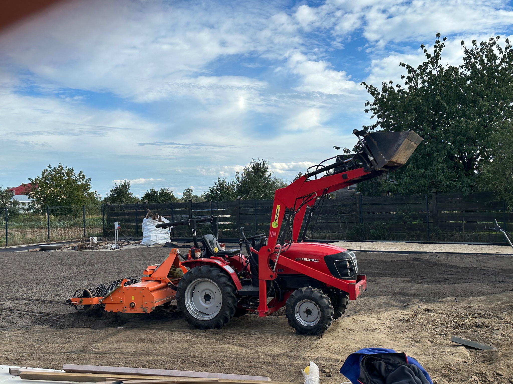 Czerwony traktor VST Fieldtrac 927 z pomarańczowym agregatem uprawowym i ładowarką czołową na przygotowanym podłożu pod trawnik, w tle worki z ziemią i ogrodzenie.