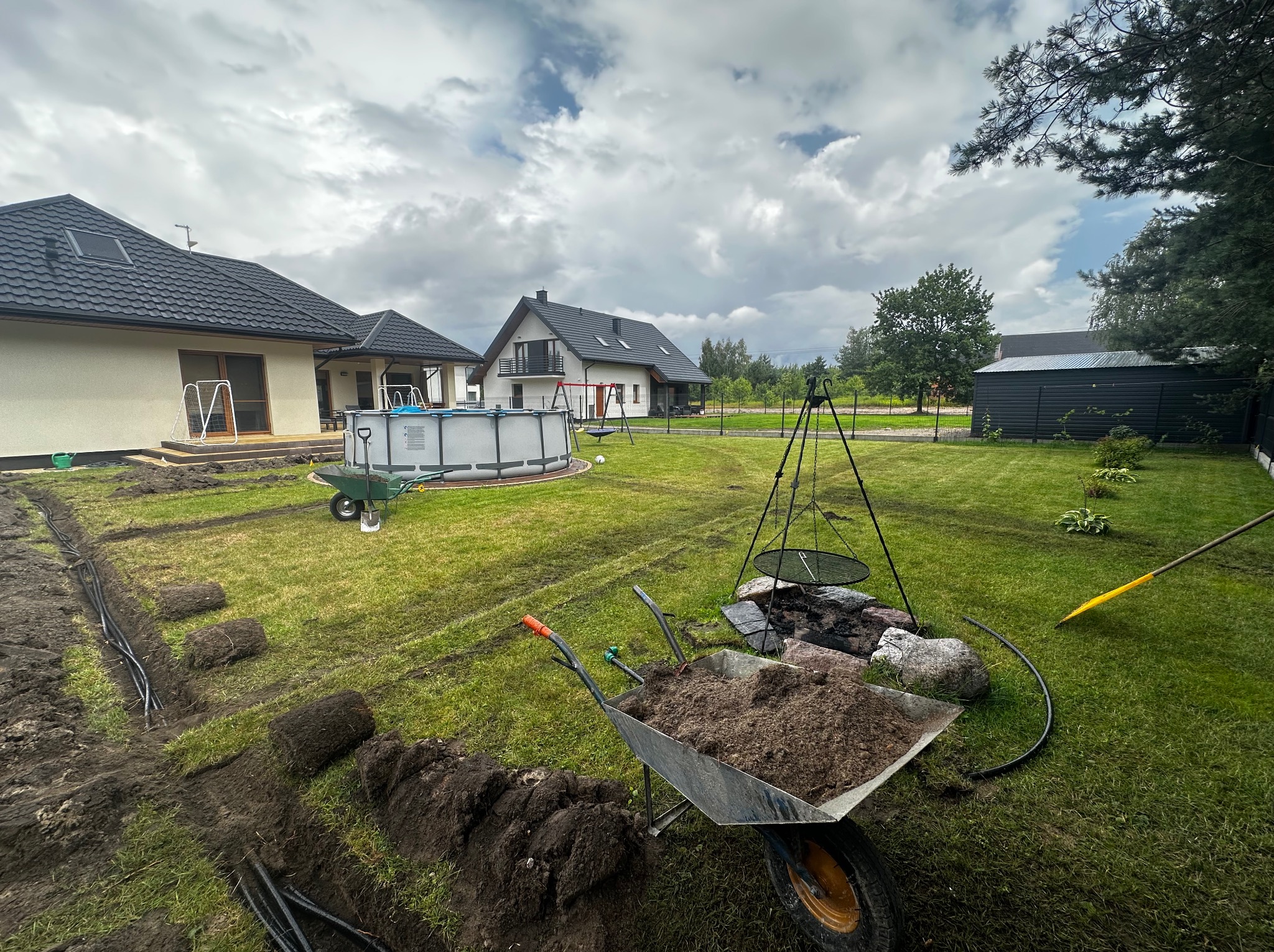 Rozpoczęte prace w ogrodzie: wykopany rów, zwinięta trawa, taczka z ziemią, widoczne domy w tle, pochmurne niebo. Przygotowania do założenia trawnika i nawadniania.