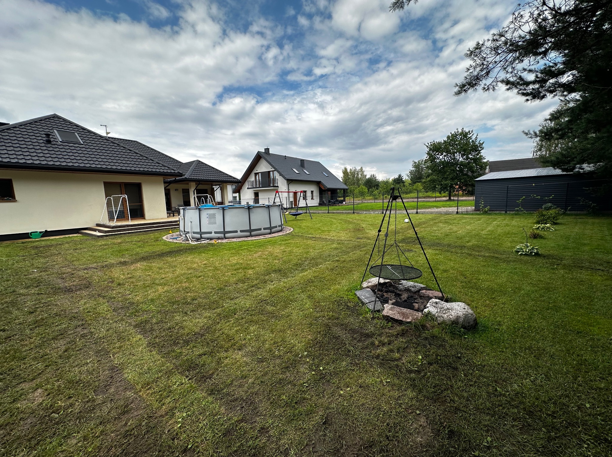 Rozległy ogród z basenem, paleniskiem i świeżo założonym trawnikiem, na którym widoczne są ślady po pracach ziemnych. W tle domy jednorodzinne i błękitne niebo.