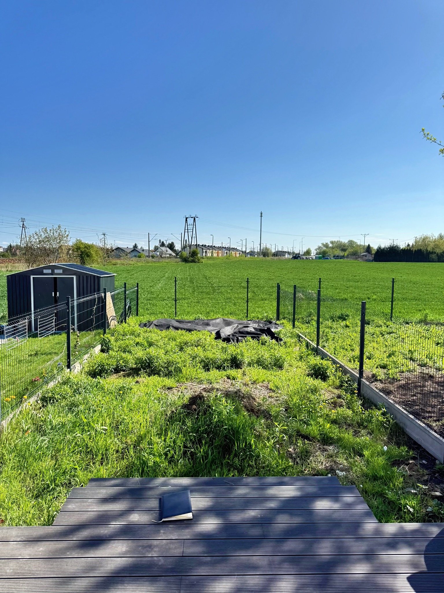 Ogród w trakcie zakładania: widok z drewnianego tarasu na teren z ogrodzeniem panelowym, przygotowywany pod uprawę. W tle widoczny blaszany domek narzędziowy.
