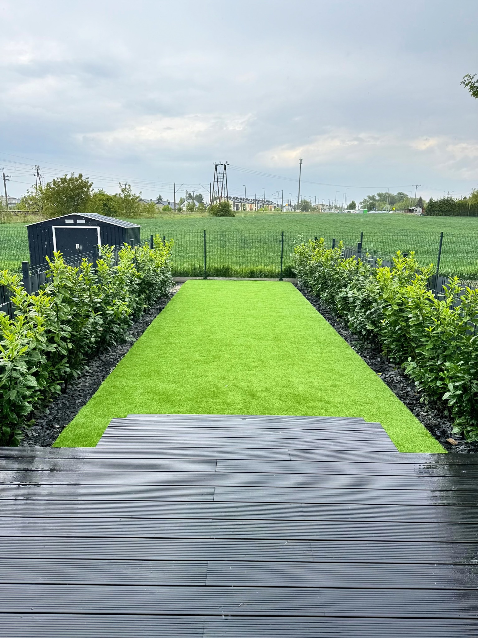 Nowoczesny ogród z równo przystrzyżonym trawnikiem, obrzeżami z kamienia, żywopłotem i tarasem z desek kompozytowych w pochmurny dzień.