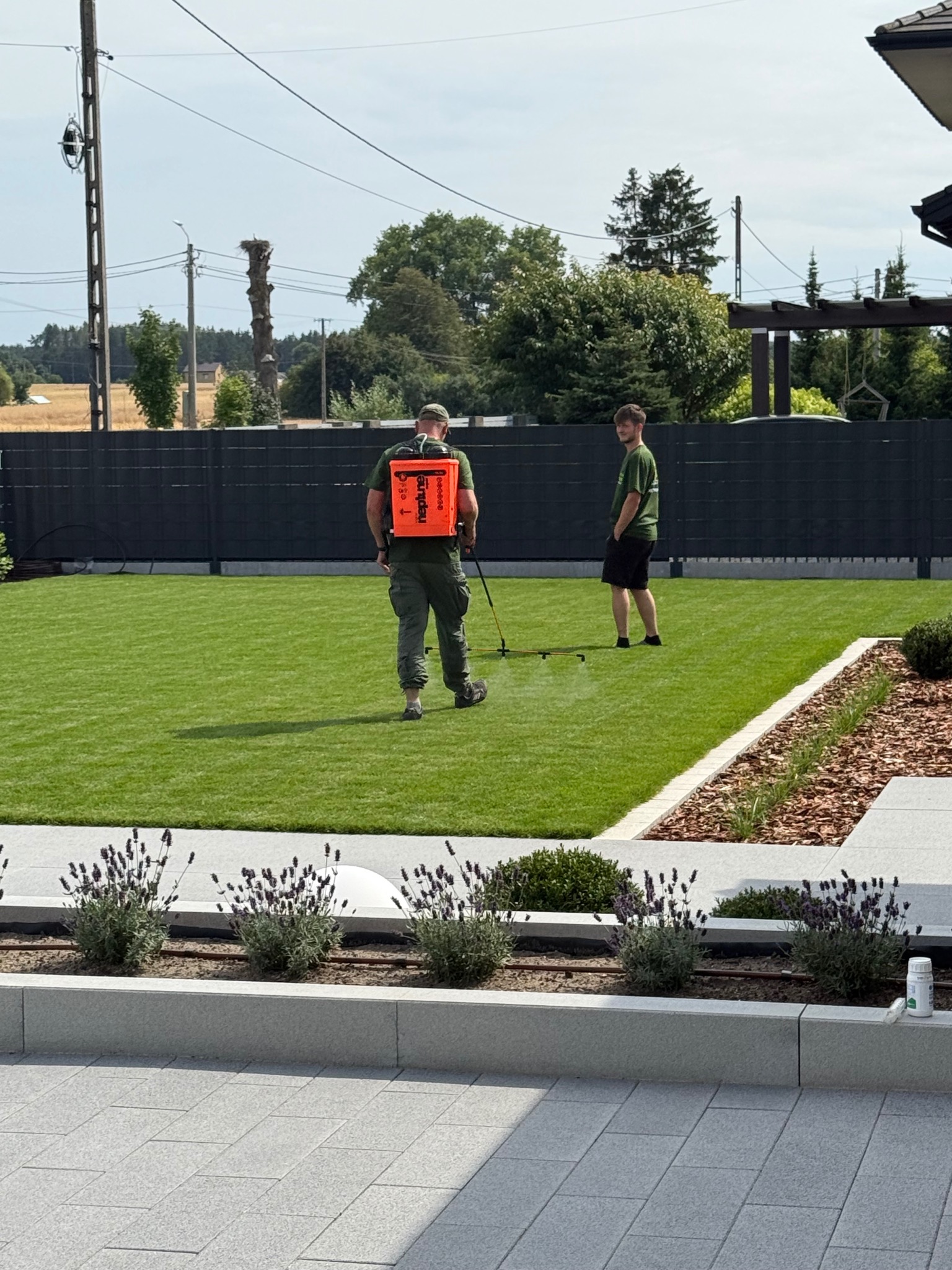 Pracownik w zielonym kombinezonie opryskuje równo przystrzyżony trawnik z użyciem opryskiwacza plecakowego. W tle drugi mężczyzna i czarne ogrodzenie panelowe.