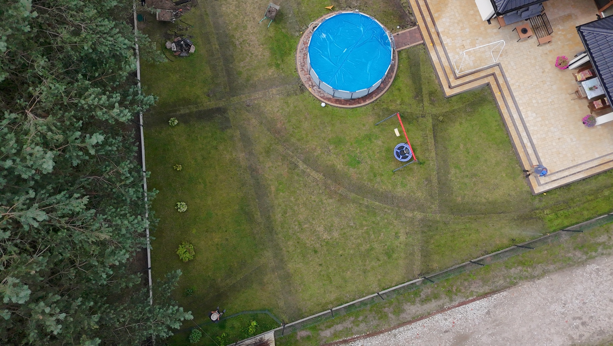 Widok z góry na zadbany ogród z basenem z niebieskim przykryciem, trampoliną, drzewami i kostką brukową przy domu. Widoczne ślady po koszeniu trawy.