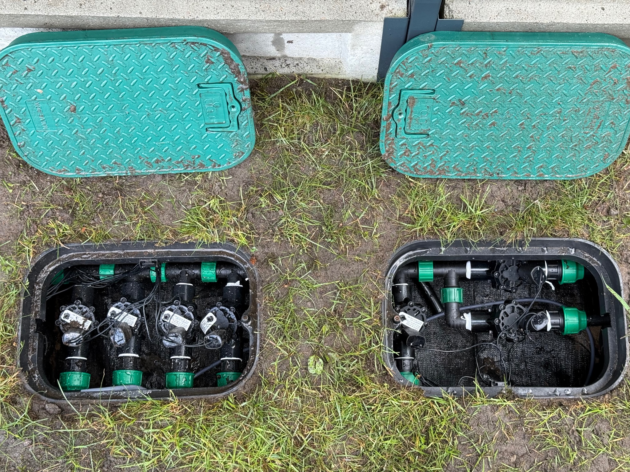 Dwa zielone, żebrowane włazy skrywające zawory nawadniające, umieszczone w ziemi z trawą. Widoczne czarne rury i złączki systemu.