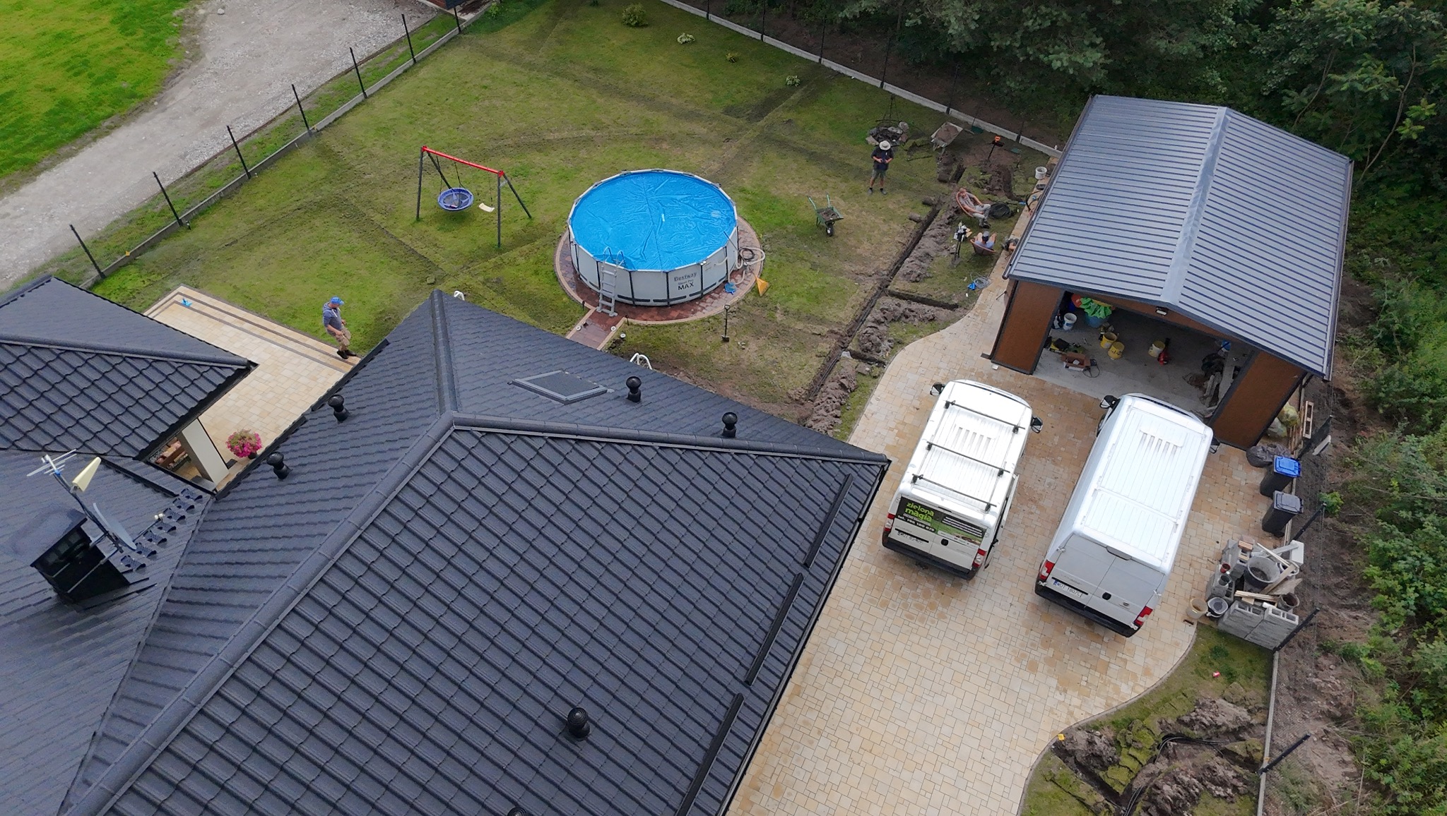 Widok z góry na dom z czarnym dachem, garaż, basen z niebieskim przykryciem, plac zabaw i dwa białe busy na posesji w trakcie prac ziemnych.