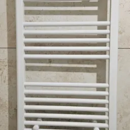 Biały, drabinkowy grzejnik łazienkowy, zamontowany na beżowej ścianie z płytek imitujących marmur, z widocznymi zaworami termostatycznymi i śladami montażu płytek u góry.