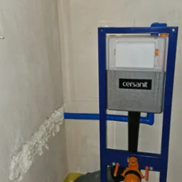 Stelaż podtynkowy Cersanit zamontowany w surowym wnętrzu łazienki, widoczne rury kanalizacyjne i instalacja wodna zabezpieczona pianką montażową.