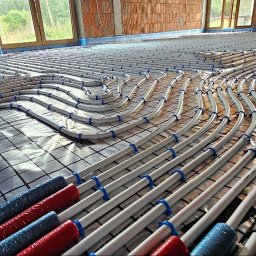 Ciepło pod kontrolą Paweł Kowalczyk - Białe rury ogrzewania podłogowego ułożone spiralnie i przymocowane niebieskimi uchwytami na folii z siatką, widoczne fragmenty czerwonych i niebieskich osłon izolacyjnych na końcach rur, surowy...