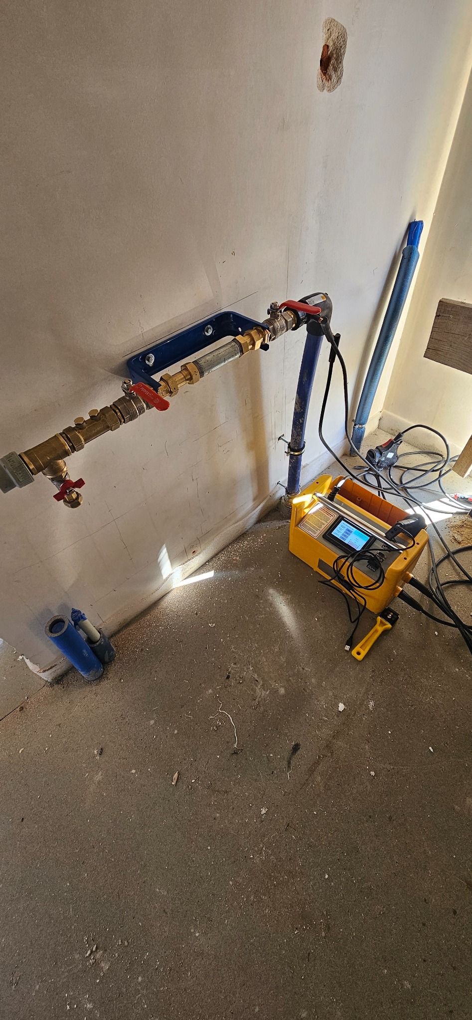 Przyłącze wodne zamontowane na ścianie, z widocznym niebieskim wspornikiem i żółtym urządzeniem pomiarowym obok, na surowej posadzce.