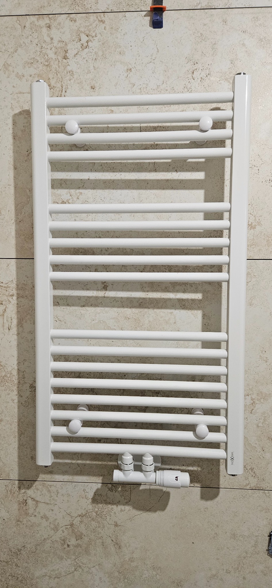 Biały, drabinkowy grzejnik łazienkowy, zamontowany na beżowej ścianie z płytek imitujących marmur, z widocznymi zaworami termostatycznymi i śladami montażu płytek u góry.