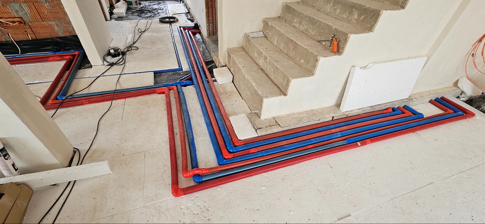 Rozłożone rury instalacji wodnej i grzewczej w kolorach czerwonym i niebieskim, na podłodze w surowym stanie, widok z góry, z fragmentem schodów w tle.
