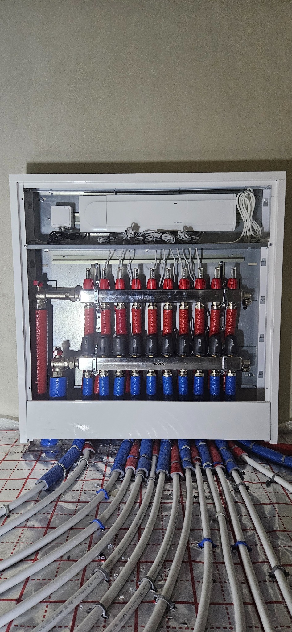 Rozdzielacz ogrzewania podłogowego Uponor w białej szafce, widoczne rury doprowadzające w izolacji czerwono-niebieskiej i sterownik elektroniczny na górze.
