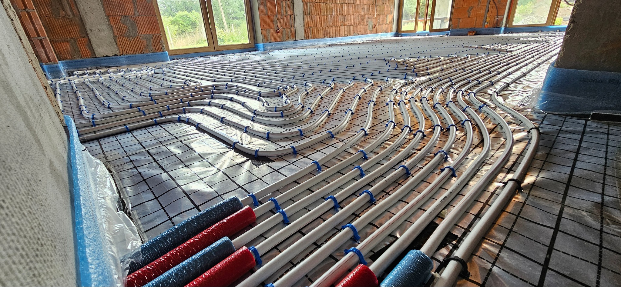 Białe rury ogrzewania podłogowego ułożone spiralnie i przymocowane niebieskimi uchwytami na folii z siatką, widoczne fragmenty czerwonych i niebieskich osłon izolacyjnych na końcach rur, surowy...