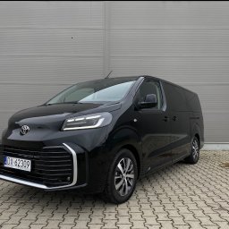 JAKUB PŁATEK LOGISTICS SOLUTIONS - Czarny van Toyota Proace Verso z polską rejestracją DX 62309, zaparkowany na brukowanej powierzchni przed szarą ścianą. Widok z przodu i z boku.