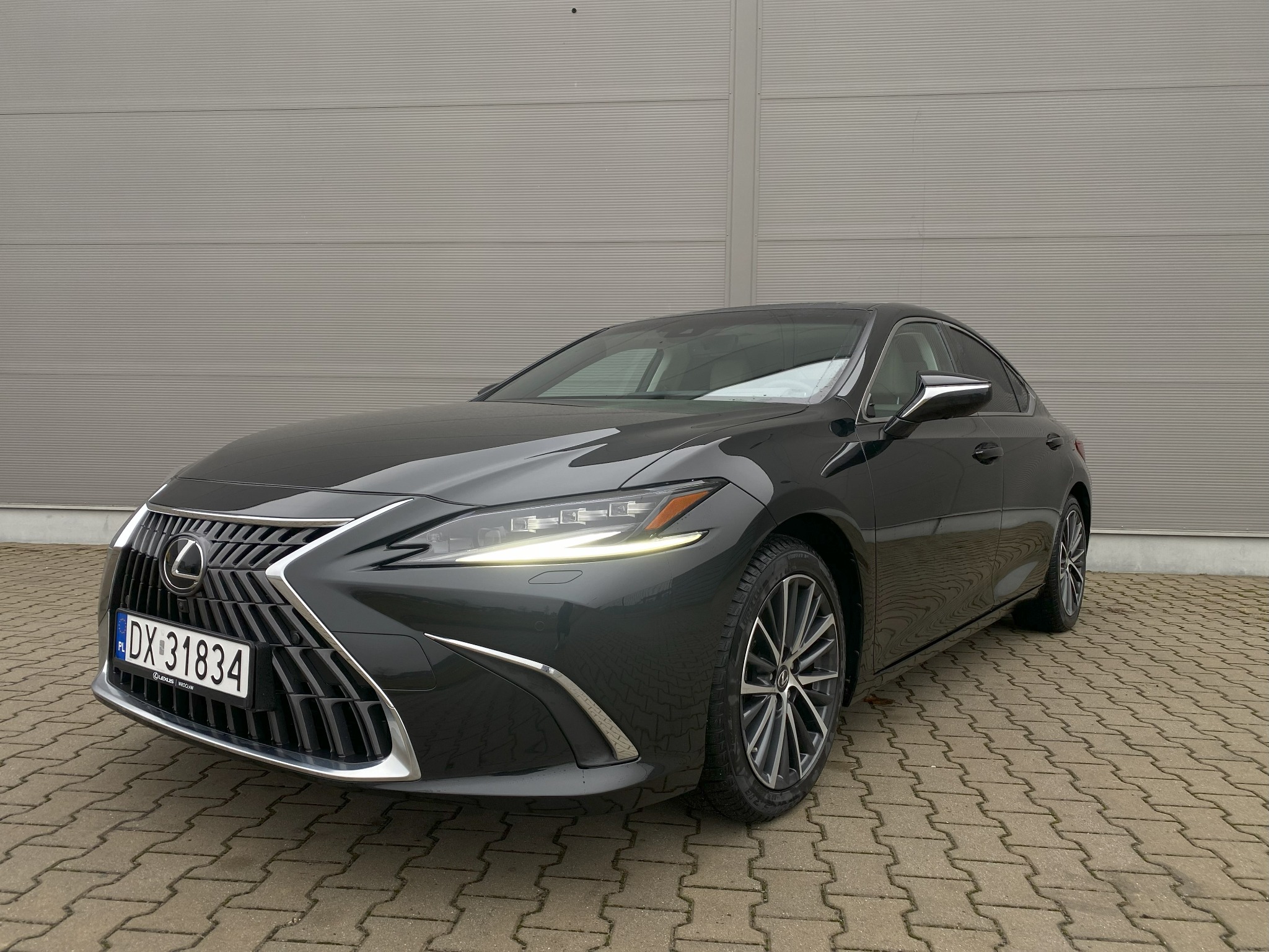 Elegancki, grafitowy Lexus ES 300h na tle szarej ściany. Widoczne detale: chromowane elementy, reflektory LED i alufelgi. Auto zaparkowane na kostce brukowej.