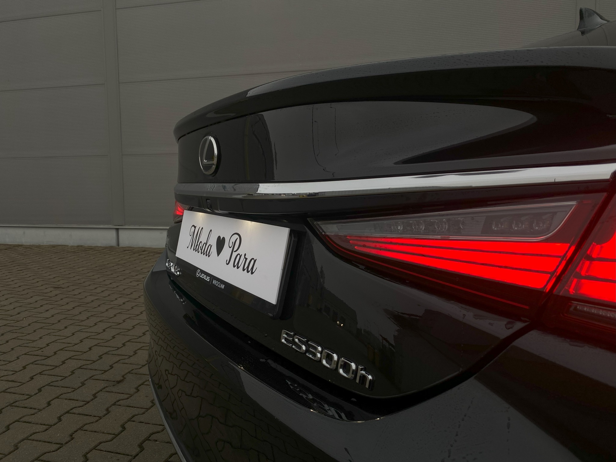 Czarny Lexus ES300h z tablicą 'Młoda Para' przy tylnej lampie, światła włączone, widok z tyłu, idealny na ślubne uroczystości.