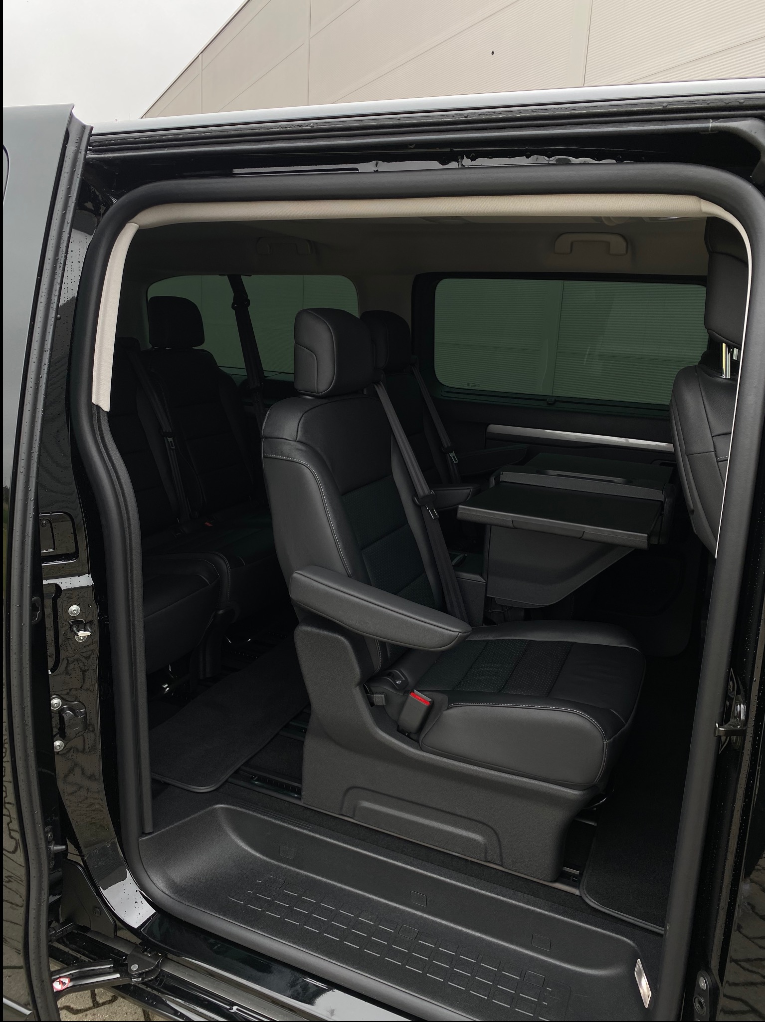 Wnętrze czarnego vana z luksusowymi skórzanymi siedzeniami, stolikami i otwartymi drzwiami, ukazujące przestrzeń pasażerską. Detaliczne wykończenie.