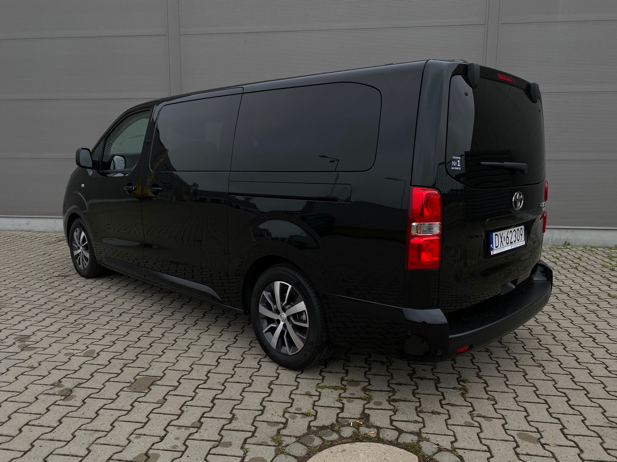 Czarny, przeszklony bus Toyota Proace Verso z tablicą rejestracyjną DX-62309, zaparkowany na kostce brukowej przed szarą ścianą. Widok z tyłu i boku.