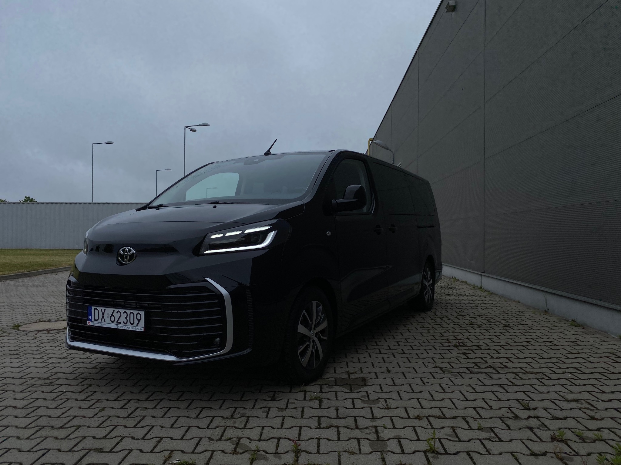 Czarny, nowoczesny van Toyota Proace Max z włączonymi światłami LED, zaparkowany na kostce brukowej przed szarą ścianą budynku w pochmurny dzień.