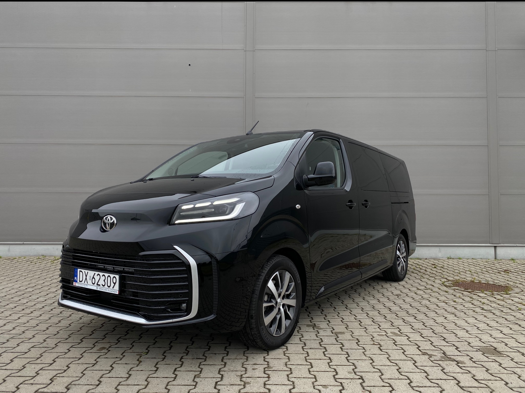 Czarny van Toyota Proace Verso z polską rejestracją DX 62309, zaparkowany na brukowanej powierzchni przed szarą ścianą. Widok z przodu i z boku.