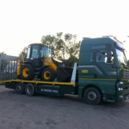 Zielona ciężar&oacute;wka z ż&oacute;łtym traktorem JCB na niskopodwoziowej platformie, widok z boku na tle drzew i drogi.