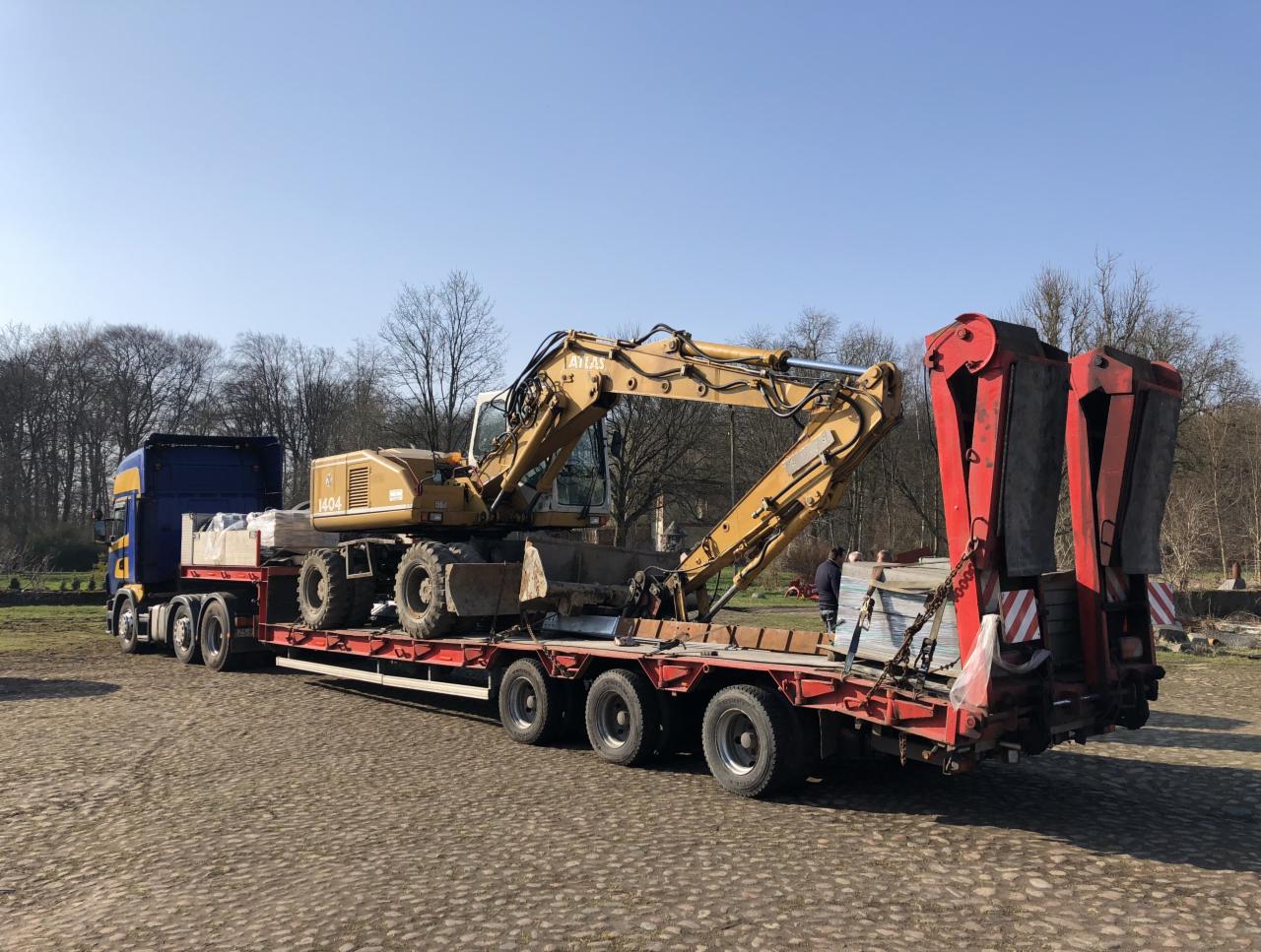 Żółta koparka Atlas 1404 przewożona na niskopodwoziowej naczepie ciągniętej przez niebiesko-żółtą ciężarówkę; z tyłu naczepy zamontowane czerwone podpory; na naczepie dodatkowo przewożone materiały...