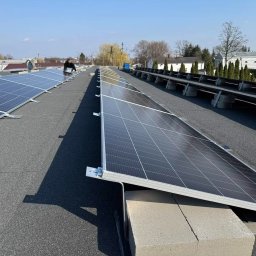 ILIOS ENERGY SPÓŁKA Z OGRANICZONĄ ODPOWIEDZIALNOŚCIĄ - Instalacja paneli słonecznych na płaskim dachu budynku z widocznym kominem w tle, pracownik w trakcie montażu.