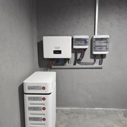 ILIOS ENERGY SPÓŁKA Z OGRANICZONĄ ODPOWIEDZIALNOŚCIĄ - Instalacja magazynu energii: białe moduły bateryjne HV ułożone pionowo, falownik ILIOS ENERGY na ścianie i skrzynki elektryczne. Szara ściana o fakturze betonu.