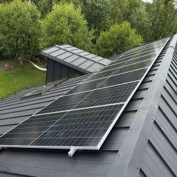 ILIOS ENERGY SPÓŁKA Z OGRANICZONĄ ODPOWIEDZIALNOŚCIĄ - Montaż paneli słonecznych na grafitowym dachu z blachy trapezowej. Widoczny pracownik z elektronarzędziem i fragment zieleni w tle.