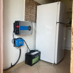 ILIOS ENERGY SPÓŁKA Z OGRANICZONĄ ODPOWIEDZIALNOŚCIĄ - Instalacja magazynu energii: akumulator, regulator ładowania i falownik na ścianie w pomieszczeniu z lodówką. Widoczne okablowanie i płytki na ścianie.