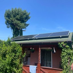 ILIOS ENERGY SPÓŁKA Z OGRANICZONĄ ODPOWIEDZIALNOŚCIĄ - Panele fotowoltaiczne na dachu domku letniskowego z zielonym dachem, na tle błękitnego nieba i drzew. Instalacja fotowoltaiczna na małej architekturze.