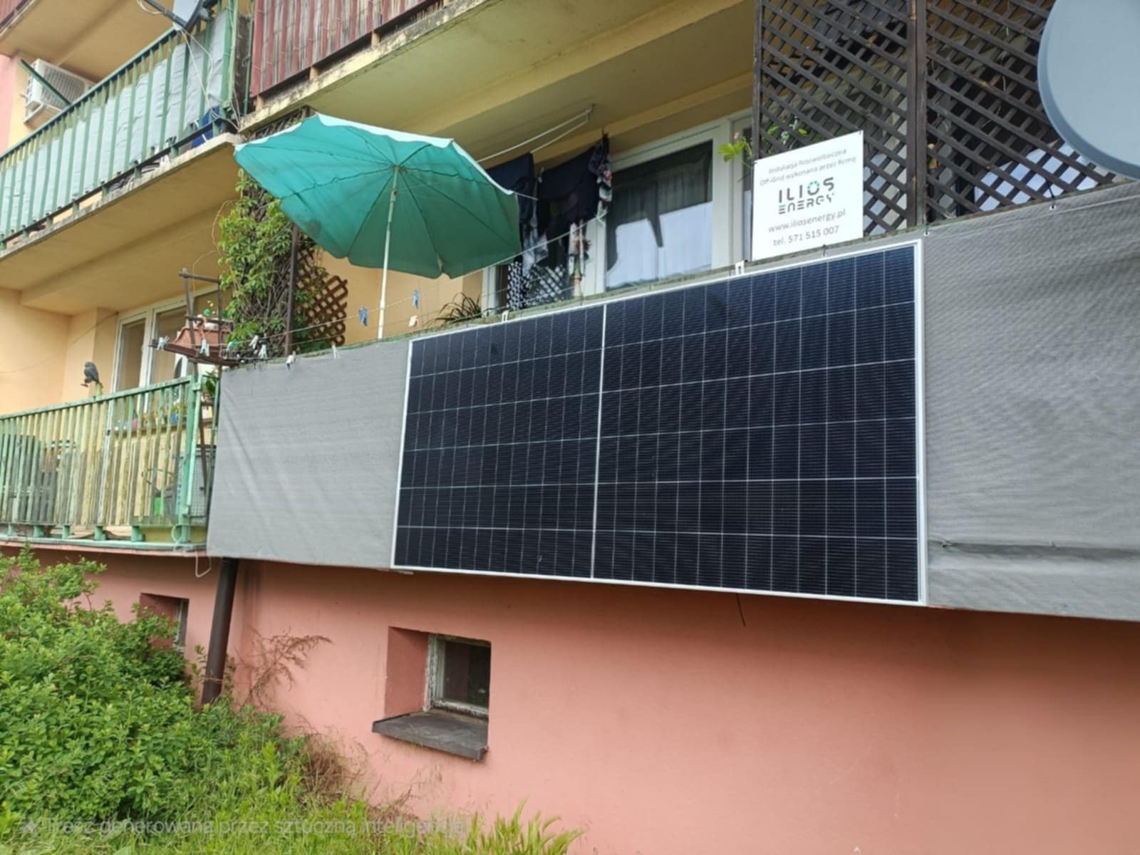 Panele fotowoltaiczne zamontowane na balkonie bloku mieszkalnego, widok z dołu na instalację. Na balkonie parasol i pranie. Tabliczka informacyjna firmy.