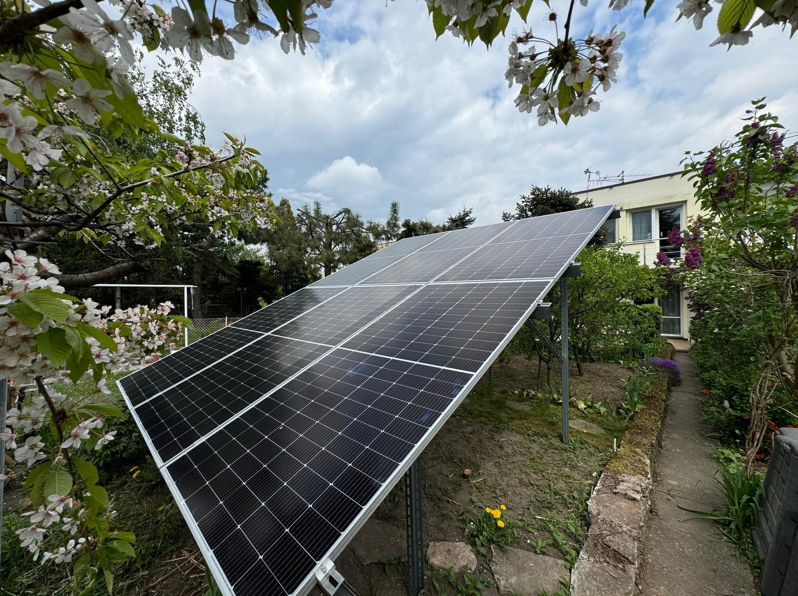 Panel fotowoltaiczny w ogrodzie, otoczony kwitnącymi drzewami i zielenią. Widoczne elementy konstrukcyjne paneli i fragment domu w tle.