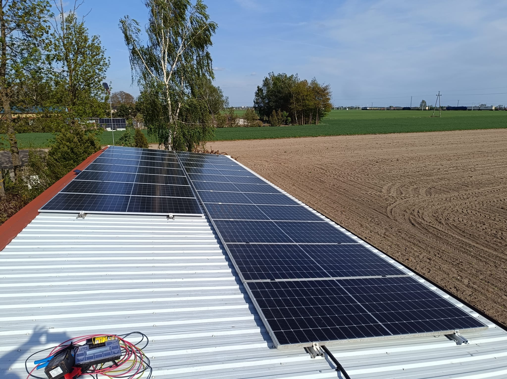 Montaż paneli słonecznych na dachu z blachy trapezowej, widok na pola uprawne i drzewa w tle. Częściowo zainstalowane panele, narzędzia pracy na dachu.