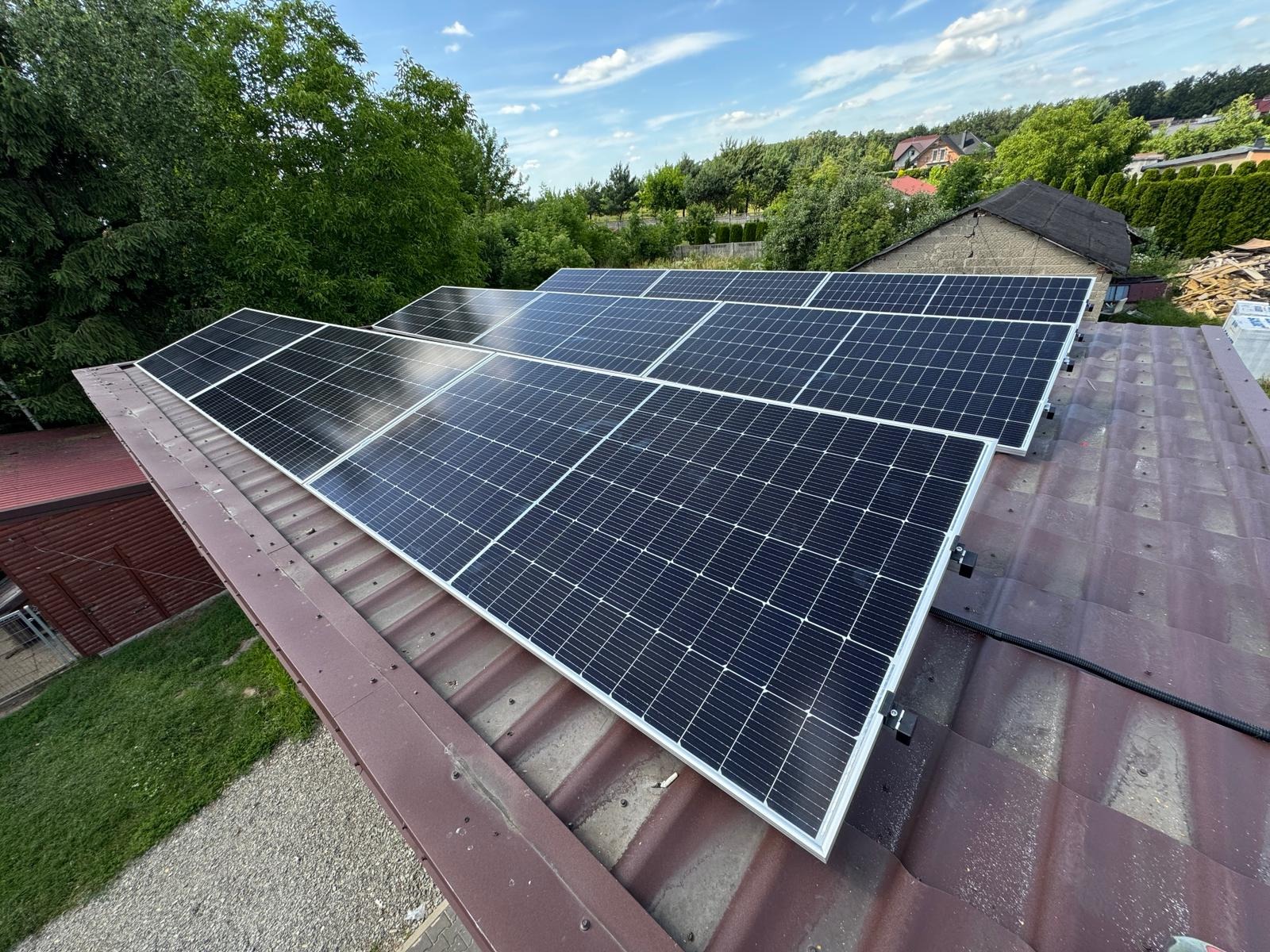 Instalacja paneli fotowoltaicznych na metalowym dachu. Widok na panele w rzędach i zielone otoczenie, błękitne niebo. Widoczne mocowania paneli do dachu.