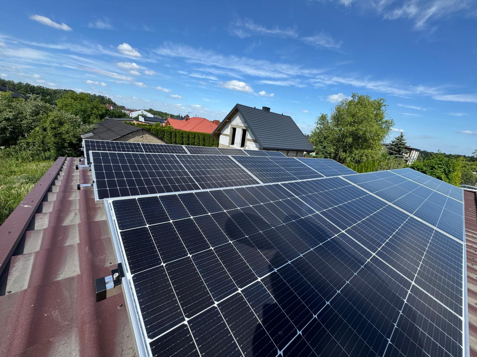 Panele fotowoltaiczne na dachu z czerwonej dachówki, w tle zabudowa jednorodzinna i błękitne niebo z chmurami.