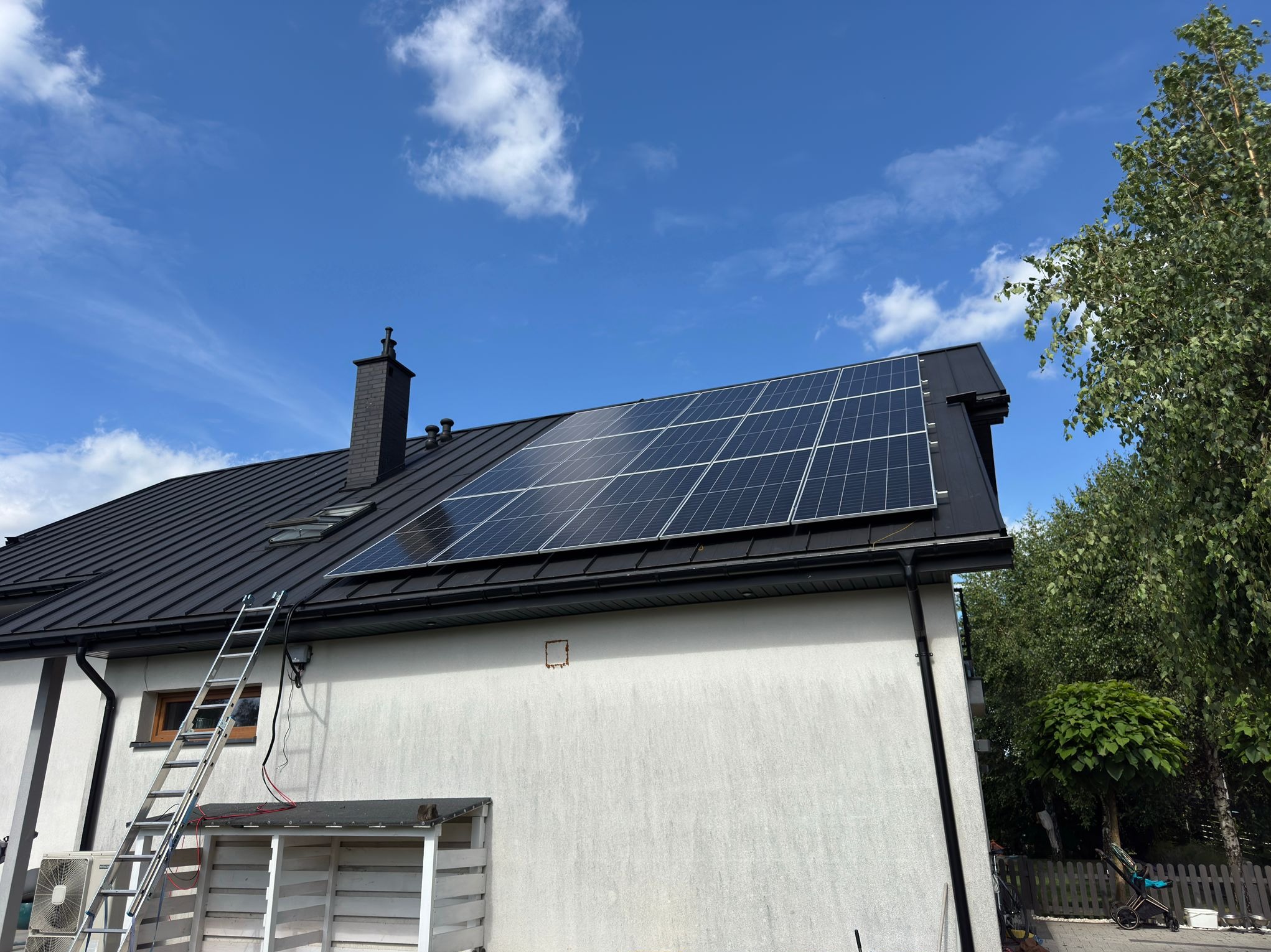 Panele fotowoltaiczne na dachu domu z czarną blachodachówką, drabina oparta o ścianę. Błękitne niebo z chmurami w tle.