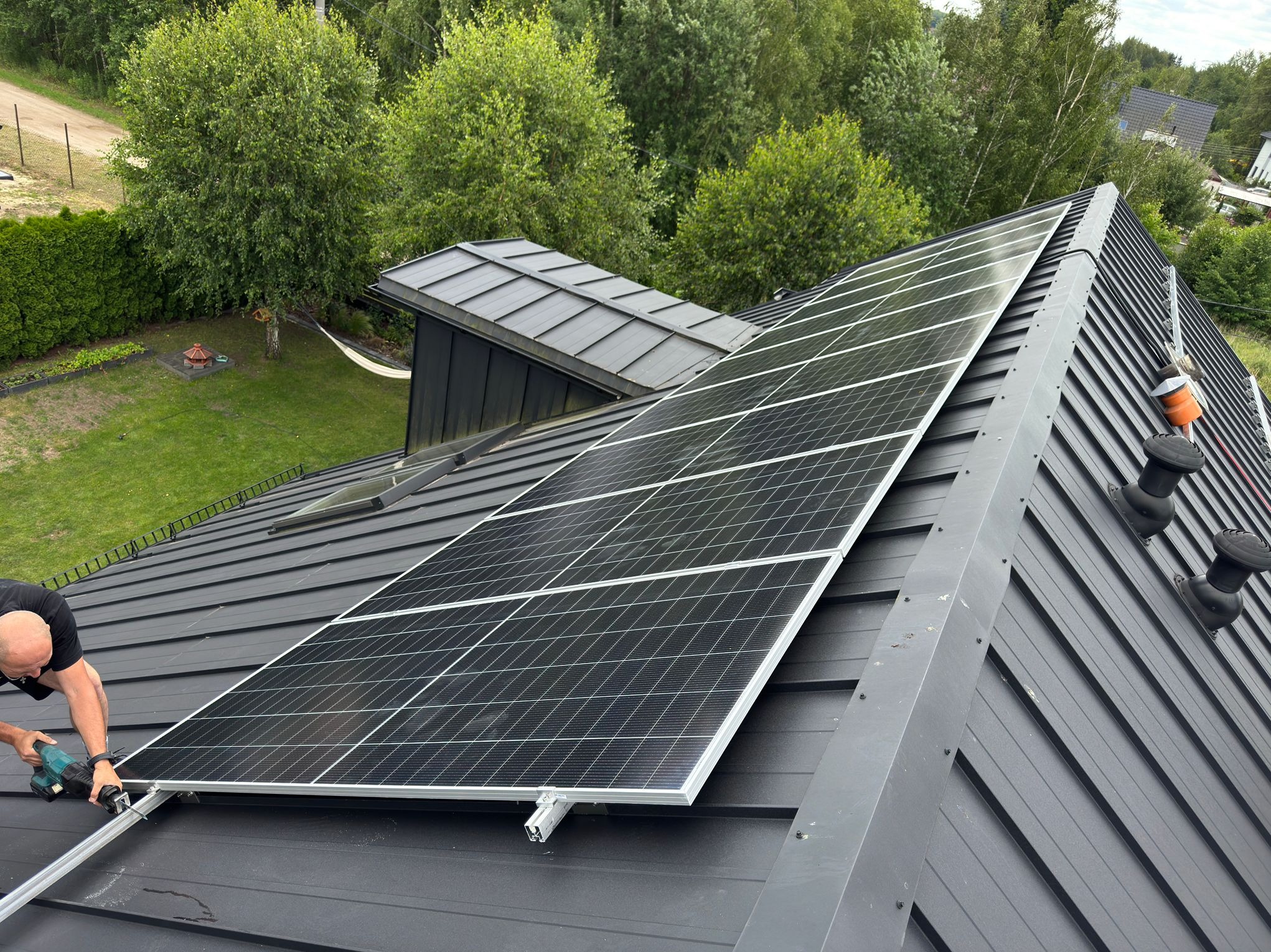 Montaż paneli słonecznych na grafitowym dachu z blachy trapezowej. Widoczny pracownik z elektronarzędziem i fragment zieleni w tle.